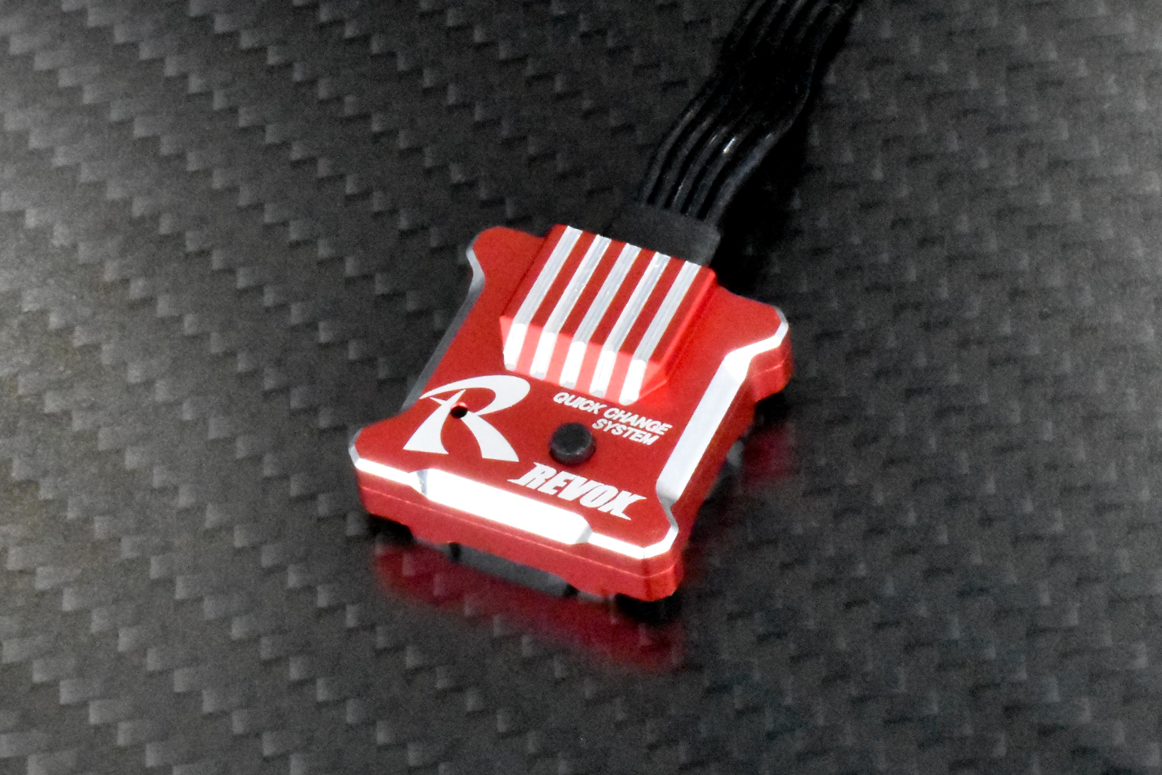 A red Rêve D REVOX drift gyro, a small anodised aluminium box with a finned top and a large R logo, sitting on a carbon-fibre surface with its black leads running out of the top of the frame.